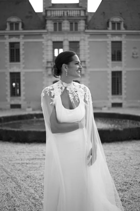 Mariée portant une longue cape brodée sur une robe fluide, devant un château