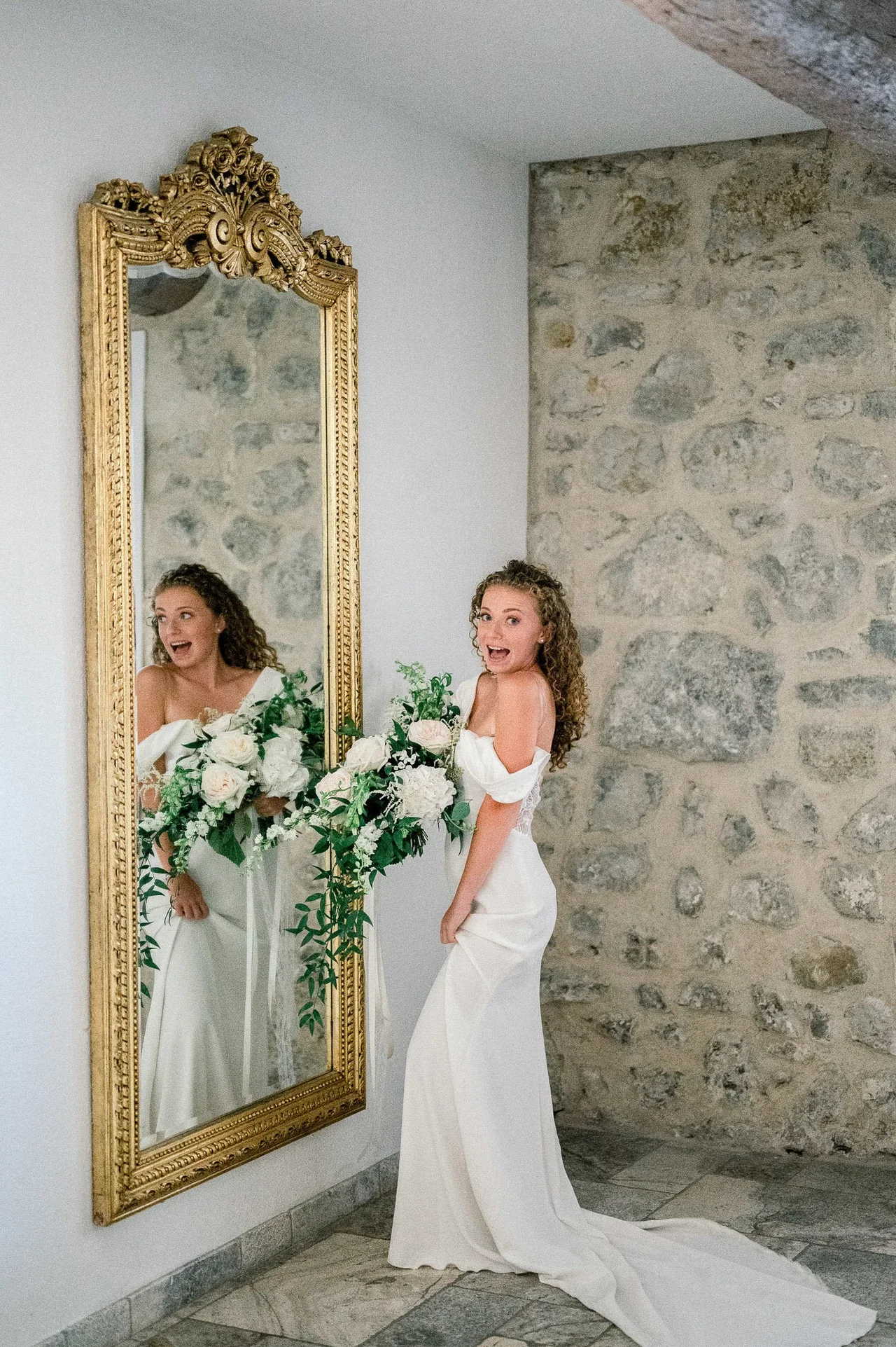 Mariée en robe épaules dénudées riant devant un grand miroir doré, bouquet blanc
