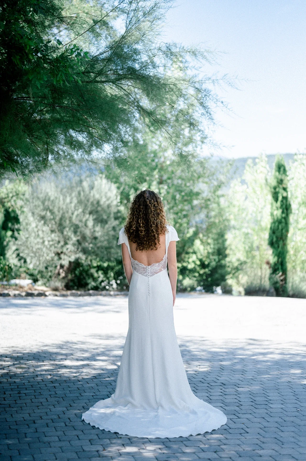 Mariée de dos en robe fourreau à dos ouvert et traîne, sous les arbres