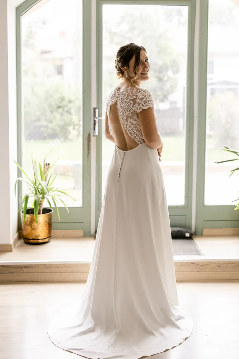 Mariée de dos en robe Lolette à dos ouvert en dentelle, près d'une baie vitrée