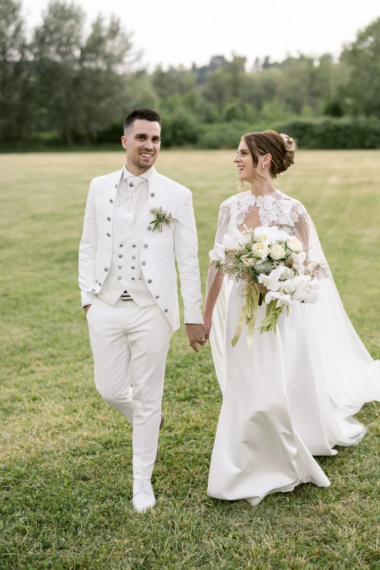 Mariés en blanc marchant dans l'herbe, cape en dentelle et grand bouquet