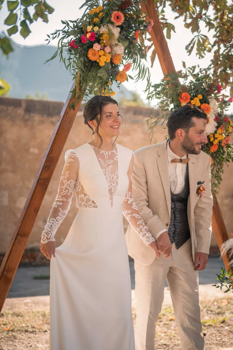 Mariés sous une arche fleurie orange, robe Lolette à manches en dentelle