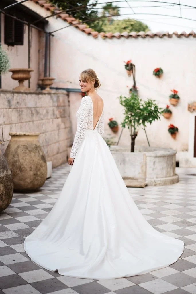 Mariée de dos dans une cour fleurie, robe à manches longues et dos ouvert