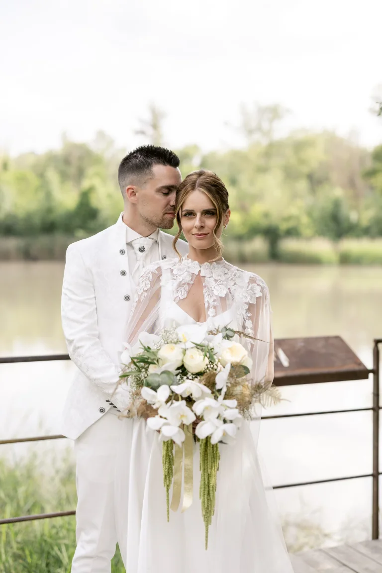 Mariés au bord de l'eau, mariée en cape de dentelle avec bouquet blanc